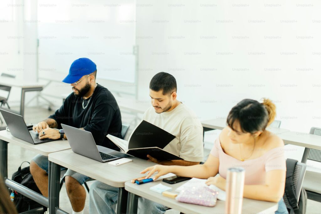 Three students in classroom using laptop, book, and writing tools for academic tasks